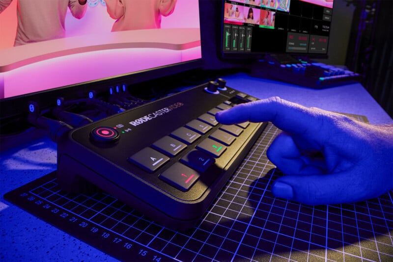 A person's hand is pointing at a button on a Rodecaster Pro II audio production console. The console is on a desk with a grid-patterned mat, and a computer monitor displaying a production interface is in the background. The scene is lit with blue and pink lighting.