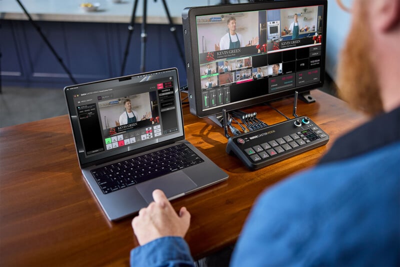 A person operates a video switcher setup including a laptop, a monitor displaying a multi-camera streaming interface, and a hardware switcher on a wooden desk. The equipment shows various camera angles of a speaker during a live broadcast or recording session.