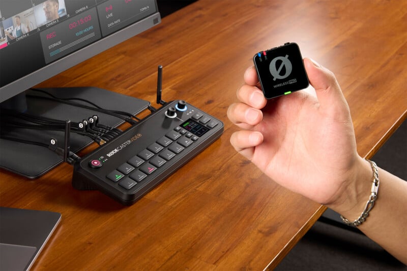 A person holds a small, square, wireless transmitter labeled "WIRELESS USB" in their left hand. On the wooden desk in front of them, there is an audio mixing console with multiple buttons, cables, and a monitor screen displaying audio settings.