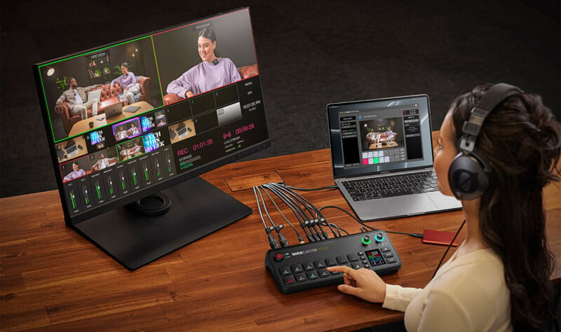 A woman wearing headphones sits at a desk in front of a monitor and a laptop, operating a video switcher with multiple cables connected. The monitor displays a multi-view screen of a video production with various camera angles showing people in a studio setting.