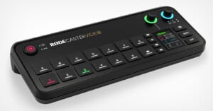 An electronic audio mixer device with the label "RODECaster Video" is shown. It features multiple black buttons, two knobs (one green and one blue), and a small digital display with various colored indicators and settings.