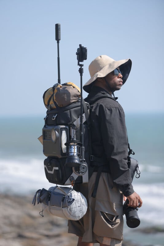 A person stands outdoors near the ocean holding a camera. They are wearing a wide-brimmed hat, sunglasses, and a backpack loaded with gear including sleeping bags, a lantern, and other equipment. The sky is clear and the ocean waves are visible in the background.