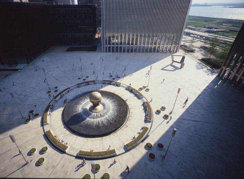 Aerial view of a large, open plaza featuring a circular fountain with a spherical centerpiece. Surrounding the plaza are modern, tall buildings. People are scattered around, and the background shows a waterfront with a few green areas. Shadows cast by the buildings cover parts of the plaza.