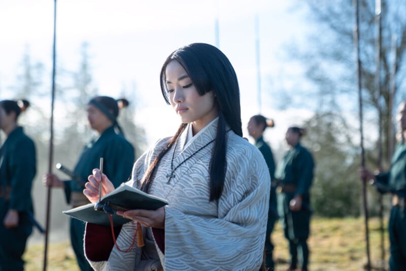 A woman in a traditional kimono stands outdoors, writing in a notebook. She is focused on her writing. Behind her, several people in blue uniforms are standing in formation holding long staffs, suggesting a training or martial arts context. Trees are visible in the background.