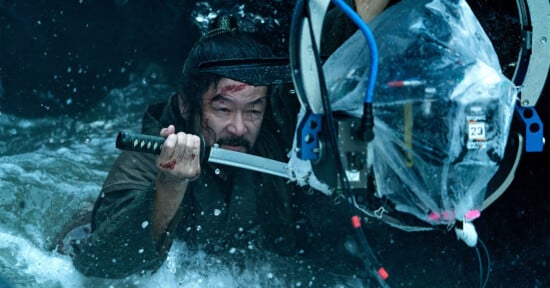 A man immersed in water grips a katana sword and appears determined. He is surrounded by a complex set of camera equipment partially covered in plastic to protect it from the water.