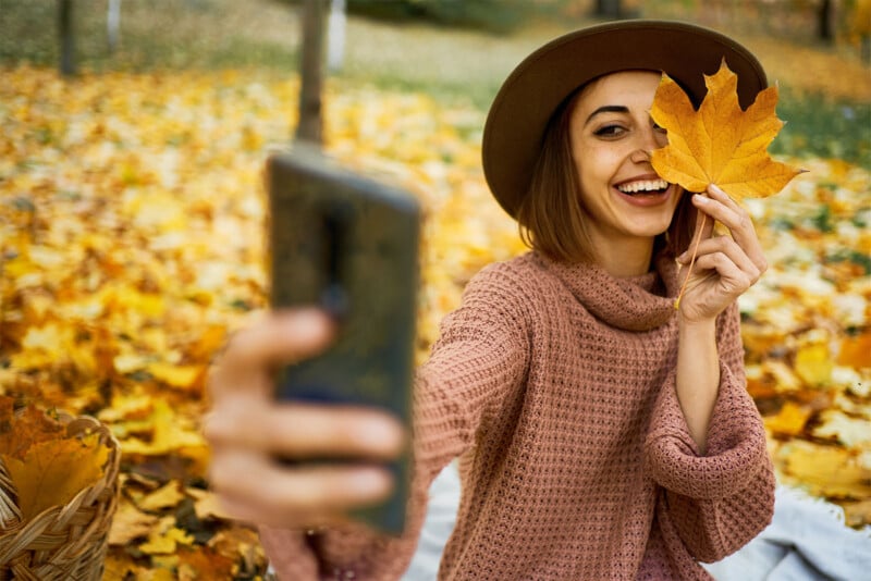 A smiling woman wearing a brown hat and a pink sweater takes a selfie while holding a large yellow maple leaf over one eye. She is sitting on a blanket in a park surrounded by fallen autumn leaves.