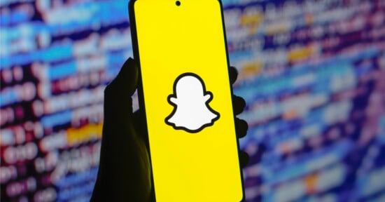 A hand holds a smartphone displaying the Snapchat logo— a white ghost on a yellow background. The background shows an out-of-focus screen filled with colorful code or text.