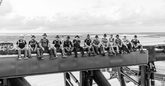 A black-and-white photo of a group of workers in hard hats and safety gear sitting on a steel beam high above the ground. The landscape in the background is flat, with clouds scattered across the sky. The workers are smiling and facing the camera.