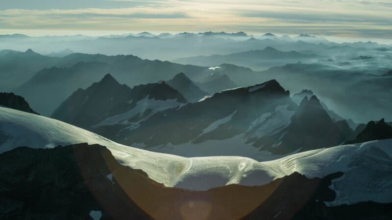 A panoramic view of a mountain range at sunrise or sunset. Snow-capped peaks and ridges create a stunning contrast with the misty valleys below. The soft light highlights the curves of the snow and casts a warm glow over the horizon, enhancing the serene atmosphere.