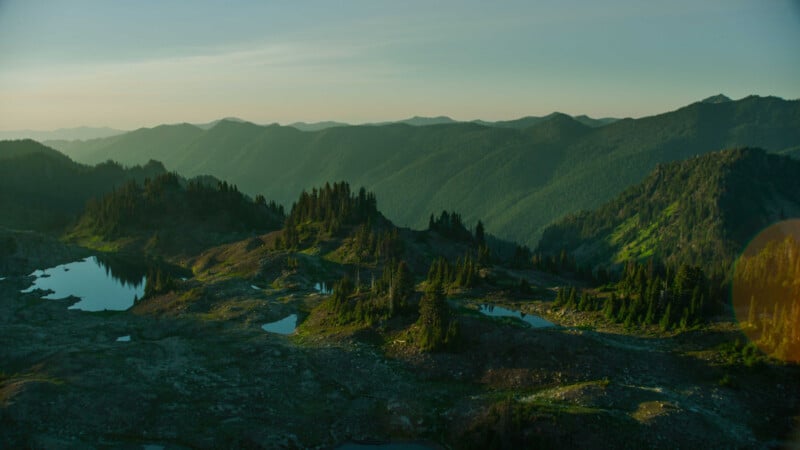 Aerial view of a mountainous landscape at dusk, featuring rolling, forested hills extending into the distance. Small reflective ponds scatter across the rocky terrain in the foreground, and the sky has a soft, warm hue from the setting sun.