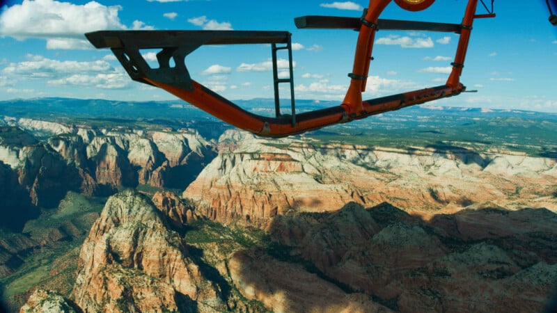 An aerial view of a vast canyon landscape with rugged cliffs and plateaus under a bright blue sky with scattered clouds. Part of a helicopter's skid is visible in the foreground, indicating the photo was taken from a helicopter.