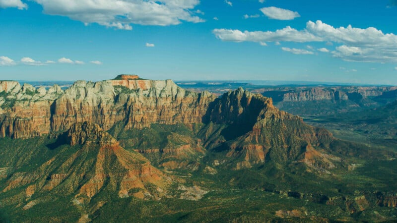 Aerial view of a vast, rugged landscape with dramatic red and white rock formations, steep cliffs, and deep valleys. The terrain is covered in patches of greenery. The sky above is clear with a few scattered clouds.