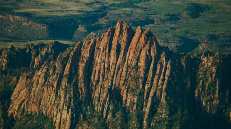 Aerial view of a striking rock formation with tall, jagged, and angular cliffs, surrounded by green vegetation and set against a backdrop of rolling hills and a distant valley. The rock's natural red and orange hues contrast with the lush greenery.