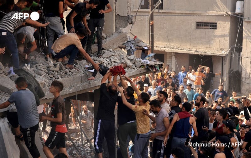 A large group of people are engaged in rescue efforts at a collapsed building site. Some individuals are on top of the rubble, pulling others up, while others provide assistance from below. Onlookers gather around the scene. The environment appears crowded and chaotic.