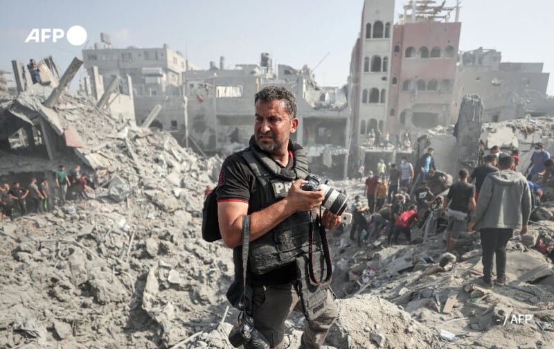 A man holding a camera stands amidst the ruins of destroyed buildings. Surrounding him are people surveying the rubble and devastation. The background shows dilapidated structures and debris scattered across the area. An "AFP" watermark is visible on the image.