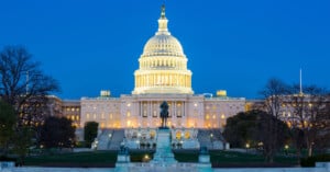 The image shows the United States Capitol building in Washington D.C. illuminated against a deep blue twilight sky. The dome and facade are lit, highlighting the architecture. A statue stands prominently in the foreground, surrounded by trees and a well-maintained lawn.