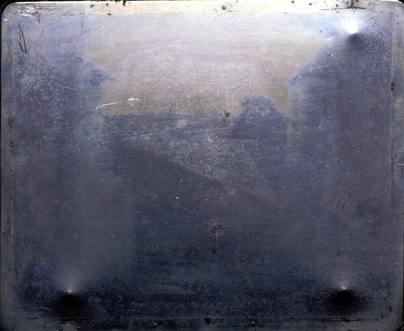 An abstract, nearly monochromatic image with a predominantly gray tone. It appears to have scratch marks, faded areas, and a textured surface, possibly metallic. The upper corners are slightly darker, and the center is somewhat lighter, giving a vague sense of depth.