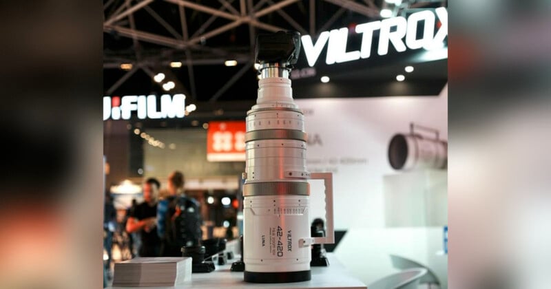 A large camera telephoto lens is prominently displayed on a table at a photography expo. The background features a booth with the FUJIFILM and VILTROX logos, as well as other photography equipment. Attendees are visible in the blurry background.