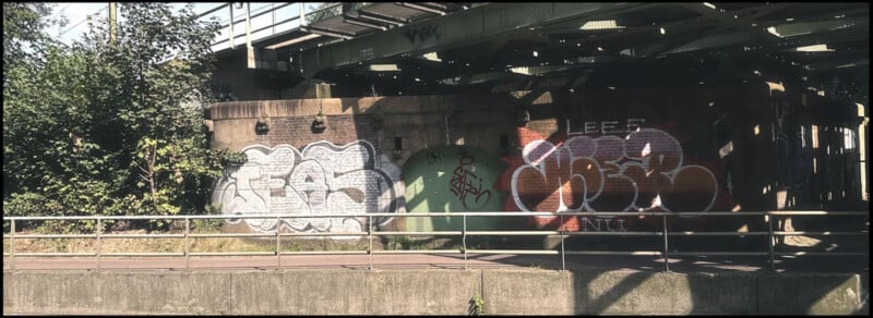 A weathered concrete and brick structure under a bridge is covered with graffiti in various styles and colors. The scene is dappled with sunlight filtering through the gaps in the bridge above. There's a metal fence in front of the graffiti and some foliage to the left.