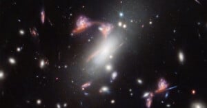 A deep space image showing numerous galaxies and stars. The central focus is a large, bright, elliptical galaxy surrounded by various other galaxies, some with distinct spiral and irregular shapes. The background is speckled with countless distant stars and galaxies.