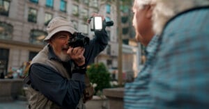 An elderly man wearing a hat and vest is taking a photograph with a camera fitted with a flash attachment. He is outdoors in an urban setting, focusing on a person in a blue shirt who is in the foreground and slightly blurred. Trees and buildings are visible in the background.