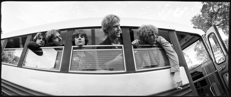 A black-and-white photo of five people sticking their heads out of the windows of a bus, looking in different directions. The bus is parked, and trees can be seen in the background. The scene captures a candid moment with a vintage feel.