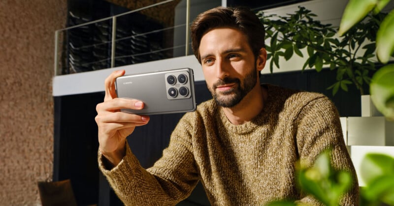 A person with a beard and short hair holds a smartphone with a sleek design and a quad camera setup on the back. They are smiling slightly and appear to be in a modern, well-lit indoor space with plants and stylish furniture.