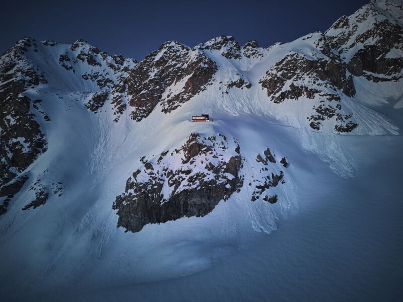 A small, illuminated cabin perched on a snowy mountain slope surrounded by rugged peaks at dusk. The snow glows softly in the fading light, creating a serene and isolated atmosphere.