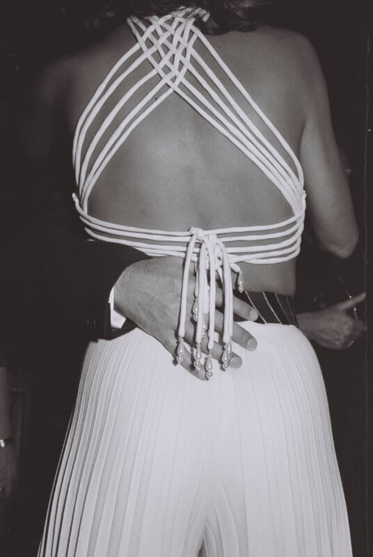 A black-and-white image of a woman in a strappy, open-back top and pleated skirt. She is being embraced by someone in a suit, whose hand rests on her lower back. The setting appears to be formal or at a social event.