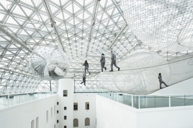 People are walking and climbing on suspended wire mesh structures inside a modern building with a high, geometric ceiling. The structures look like netted tubes and bubbles, and there are several floors visible below.