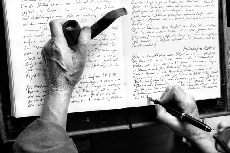 A person holds a pipe in one hand while writing in a notebook with the other hand. The notebook is filled with handwritten text. The image is in black and white, highlighting the details and texture of the hands and writing.