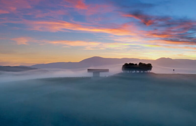 A serene landscape at dawn with a foggy field, silhouetted trees, and a building in the foreground. The sky is painted with hues of pink, orange, and blue, with distant mountains visible under the colorful sky.