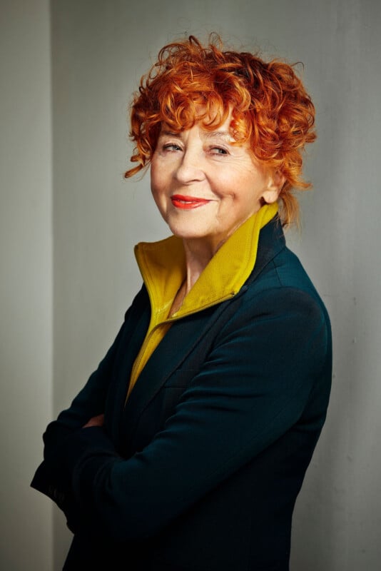 An older woman with curly red hair and a bright yellow shirt stands against a plain wall, smiling confidently with her arms crossed. She is wearing a dark blazer.