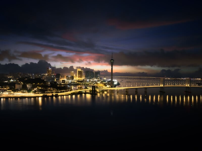 A scenic nighttime view of a cityscape with illuminated buildings, a tall tower, and a bridge reflecting on the calm water. The sky displays a gradient of deep blues and crimson, suggesting a sunset or pre-dawn setting.