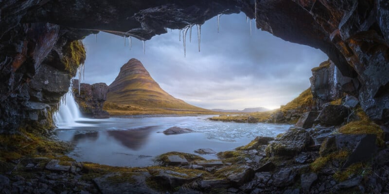 A stunning vista from inside a cave, looking out toward a iconic mountain with a conical shape. A cascade flows into a calm river, creating a serene scene. Icicles hang from the cave's roof, adding to the mystical atmosphere. The sky is overcast, casting a soft light.