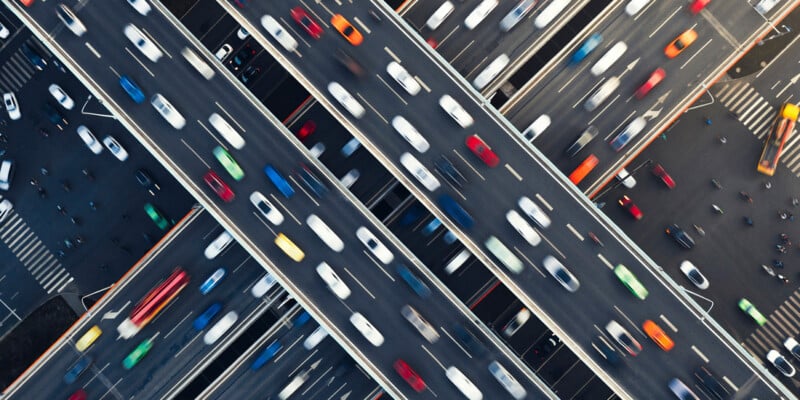 Aerial view of a busy multi-lane highway intersection with several overpasses and numerous vehicles. The cars and trucks are moving in various directions, creating a dynamic and bustling scene indicative of urban traffic. The roadways are mostly parallel and intersecting.