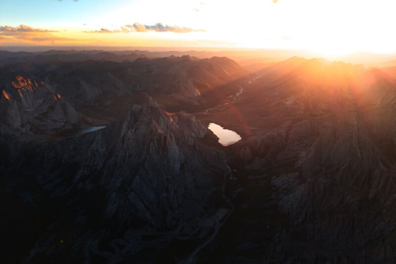 A breathtaking mountain landscape at sunset, with a rugged terrain of sharp peaks and valleys. The sun casts a golden glow over the horizon, illuminating a serene, reflective lake nestled among the jagged mountains.