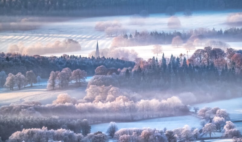 A serene winter landscape featuring frosted fields and trees under a soft mist. A church steeple rises above the treetops in the distance, surrounded by a quiet forest, under a pale blue sky.