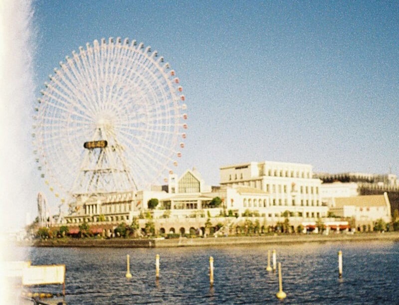 A scenic waterfront view features a large Ferris wheel with colorful gondolas, situated near a complex of light-colored buildings. The image captures a clear blue sky and calm water with wooden posts in the foreground.