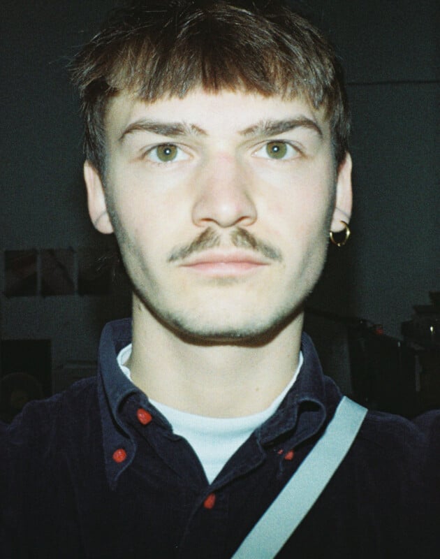 A person with short hair, a mustache, and an earring looks directly at the camera in a dimly lit room. They are wearing a dark shirt with a white collar visible underneath.