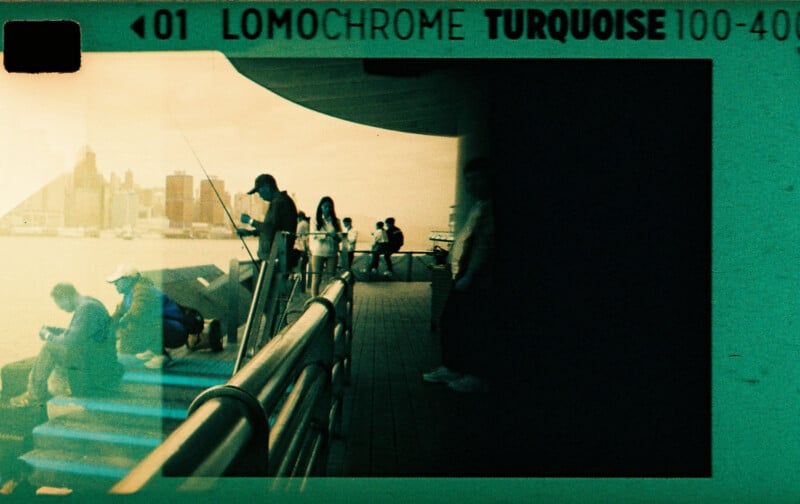 People stand and sit along a waterfront railing, some fishing, against a city skyline. The image has a vintage, teal tint with film details visible at the top.