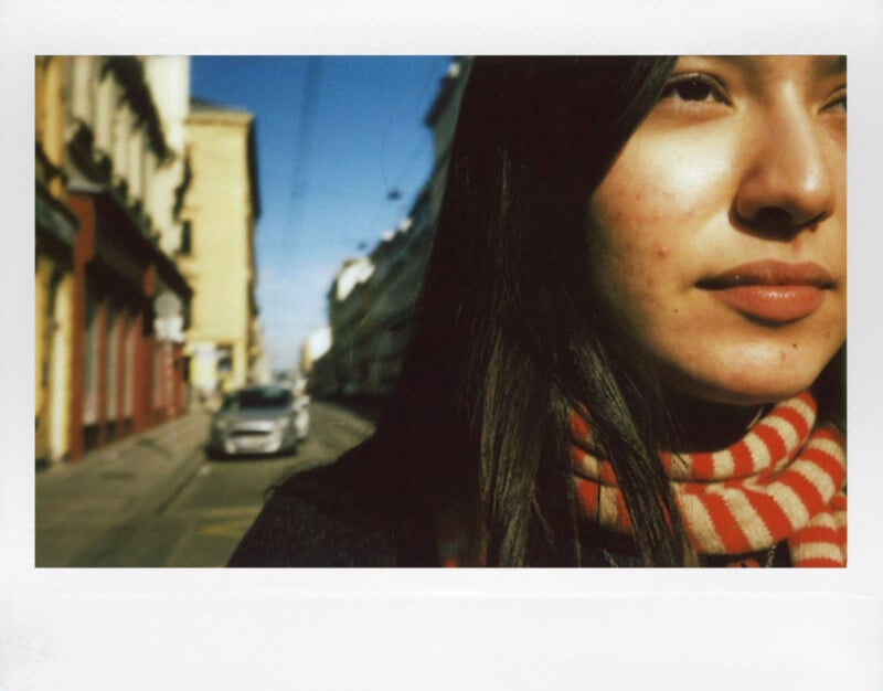 A person with long dark hair and a red striped scarf stands on a city street. The background shows a car and buildings under a clear blue sky. The focus is on the person's face, capturing natural light and shadows.