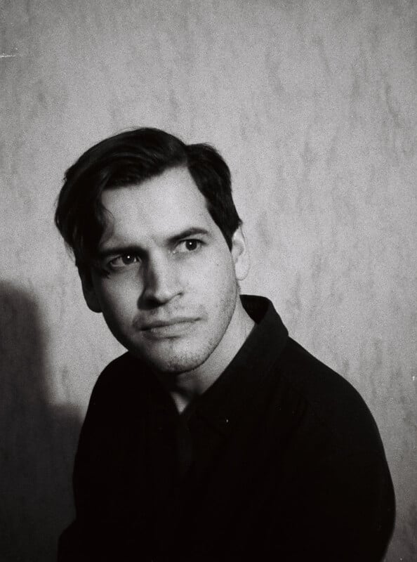 A black and white portrait of a person with short, dark hair, wearing a dark shirt. They are looking slightly to the side with a thoughtful expression. The background is plain.