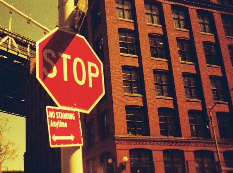 A vintage-style photo showing a red stop sign and a "No Standing Anytime" sign on a post, with a large brick building in the background. The sky is clear, and part of a bridge can be seen on the left.