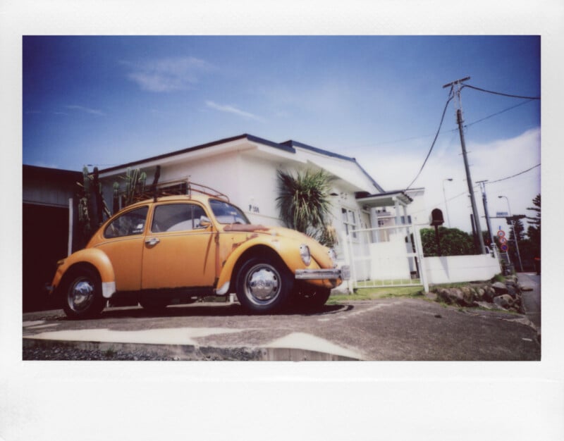 A vintage orange Volkswagen Beetle with a roof rack is parked in front of a white house. The sky is clear and blue, and the scene captures a sunny day with palm trees and utility poles in the background.