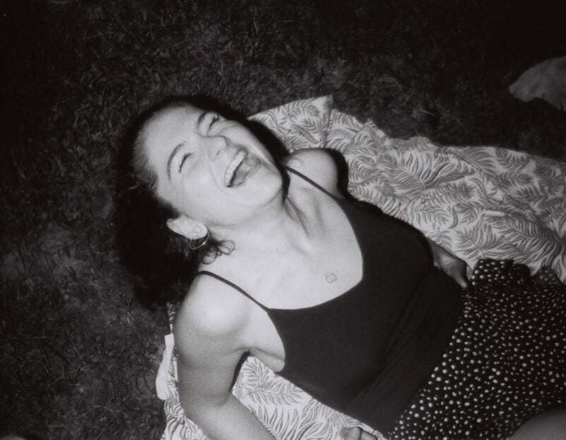 A black-and-white photo of a person lying on a patterned blanket outdoors, laughing joyfully. They are wearing a black tank top and dotted pants, with their head tilted back and eyes closed.