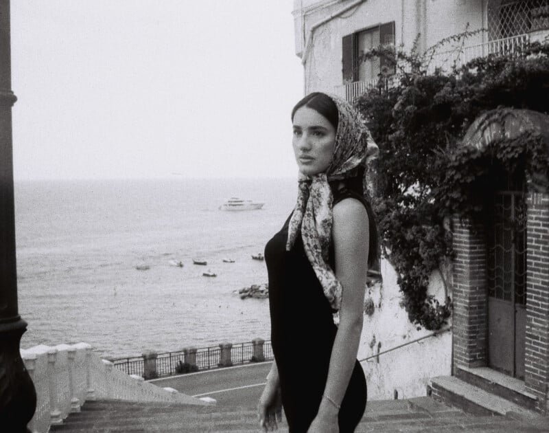 A woman wearing a floral headscarf and sleeveless dress stands on a staircase overlooking the sea. Boats are visible on the water, and buildings with ivy-covered walls appear to the right. The scene has a vintage, black-and-white aesthetic.