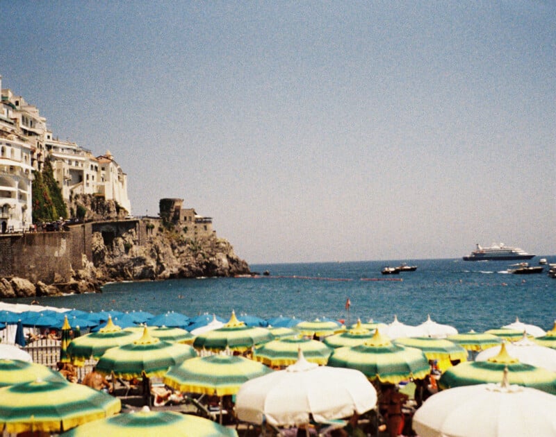 A picturesque seaside view with bright yellow and green beach umbrellas covering the sandy shore. In the distance, the blue sea stretches out with boats sailing, and a hillside town with buildings perched atop a rocky cliff.