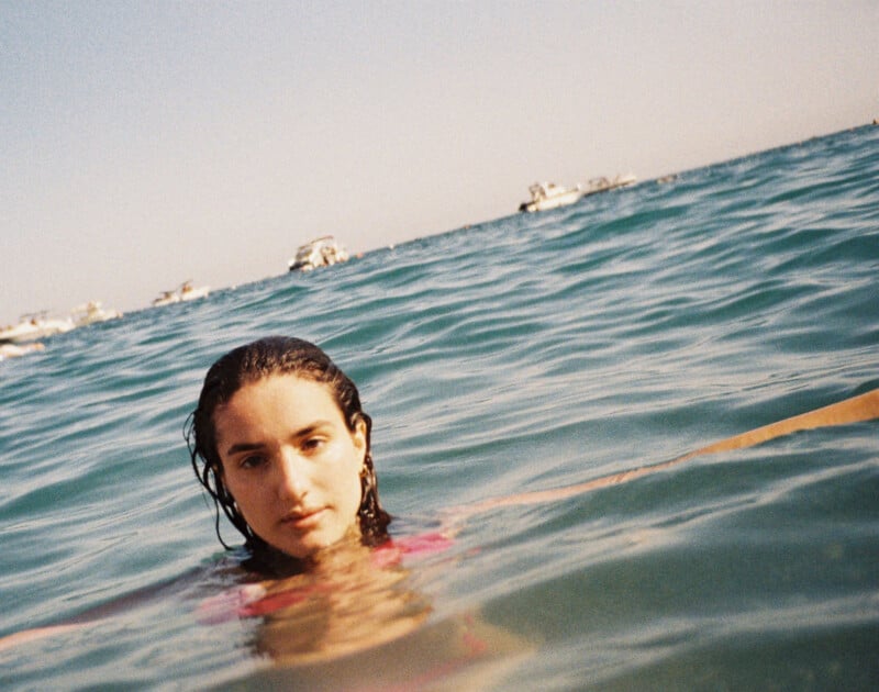 A person with wet hair is partially submerged in clear blue water, looking towards the camera. The horizon is visible in the background, with several boats scattered across the ocean under a clear sky.