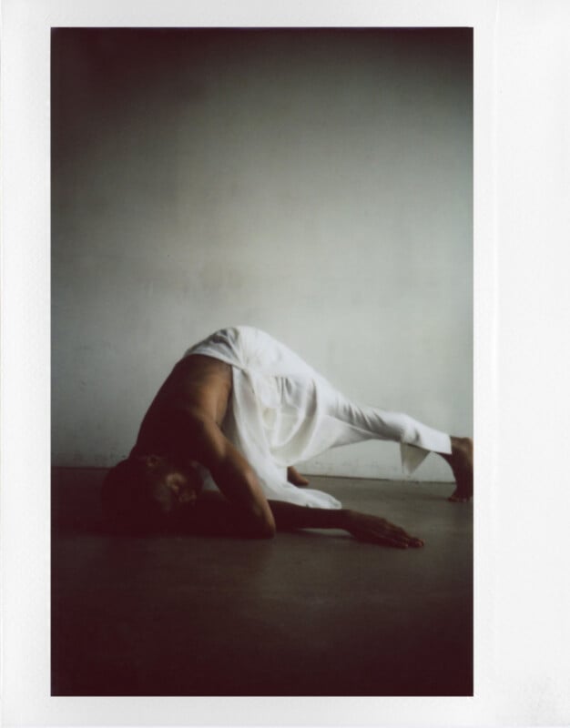 A person wearing a white outfit performs a graceful dance move, arching back with one arm touching the floor. The minimalist setting features a plain, slightly shadowed wall, creating a serene and contemplative atmosphere.